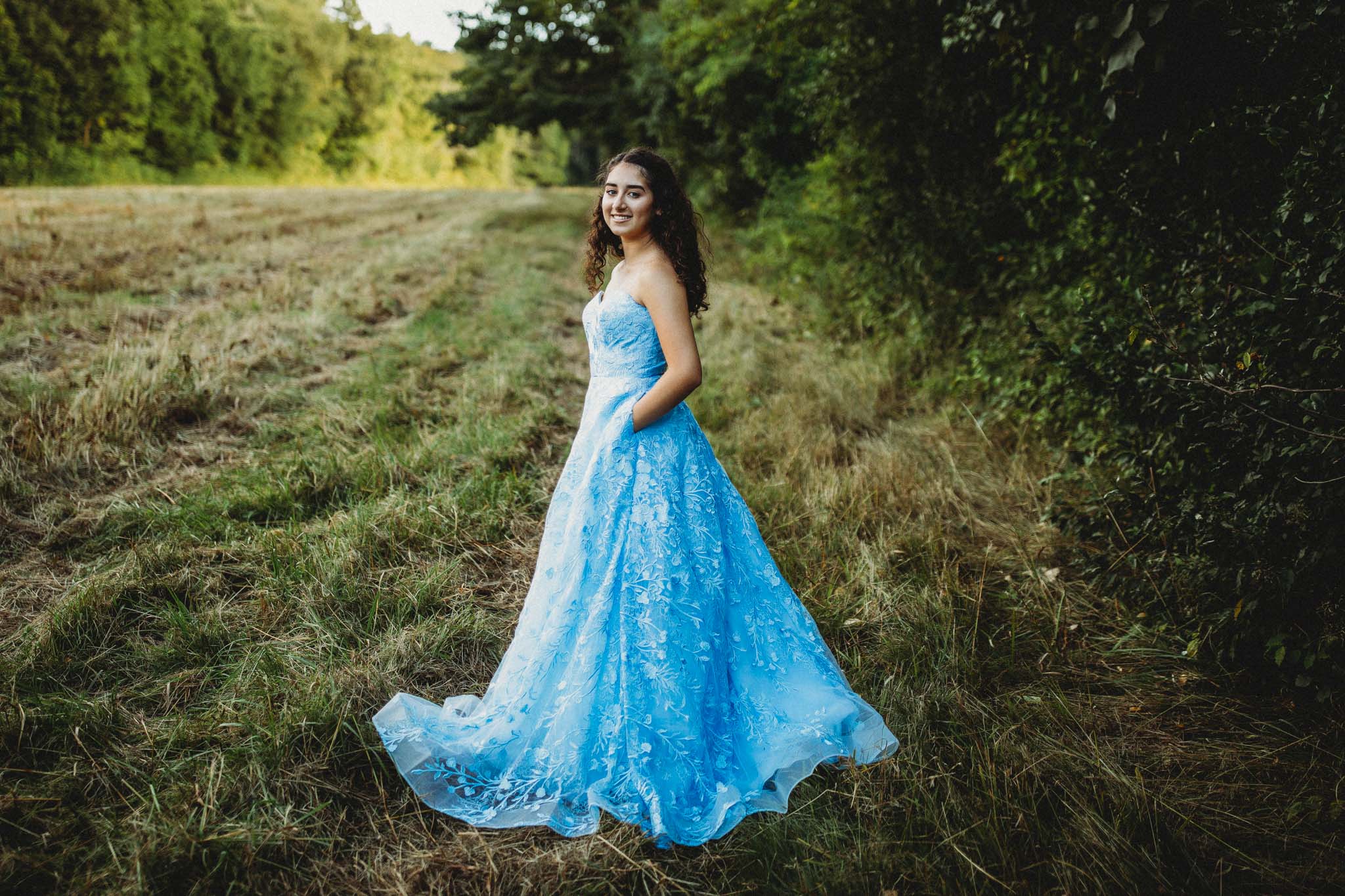 Senior portrait of a young woman wearing a long blue strapless prom dress with a floral pattern while standing in a field of long grass in Massachusetts