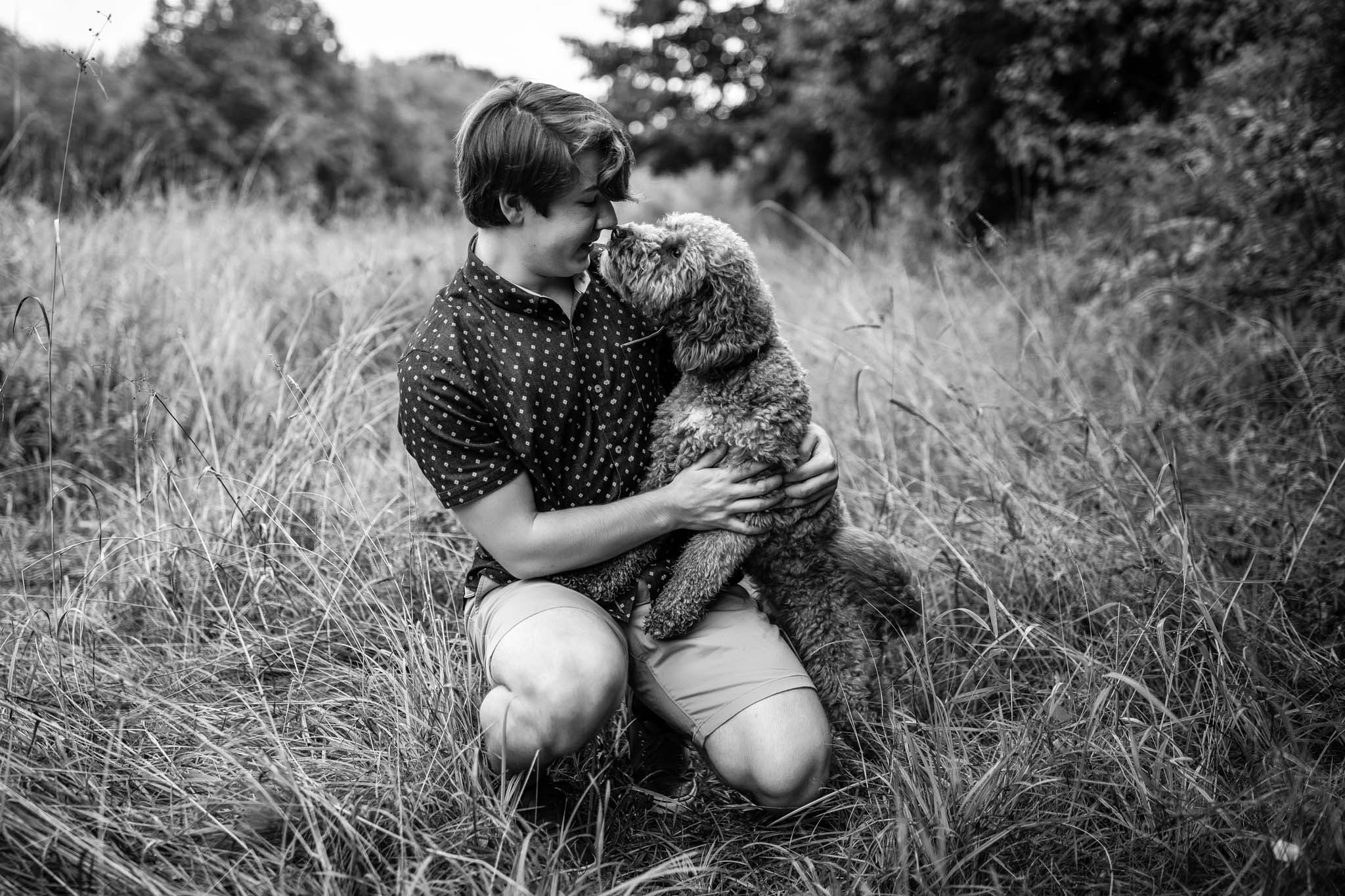 Black and white high school senior portrait of a young man kneeling with his dog in a field of long grass and trees in Massachusetts