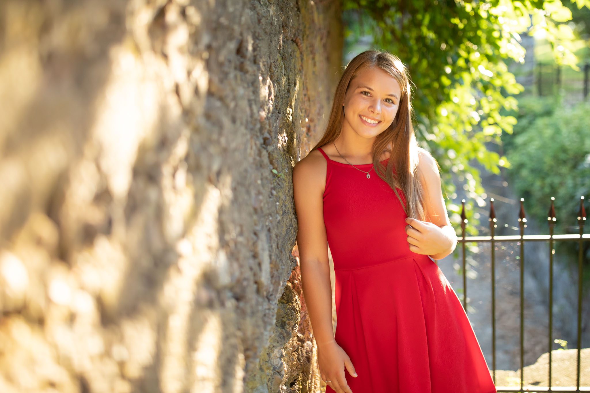 Summer Senior portrait of a young woman in a casual red dress at sunset while leaning against a stone wall in Massachusetts