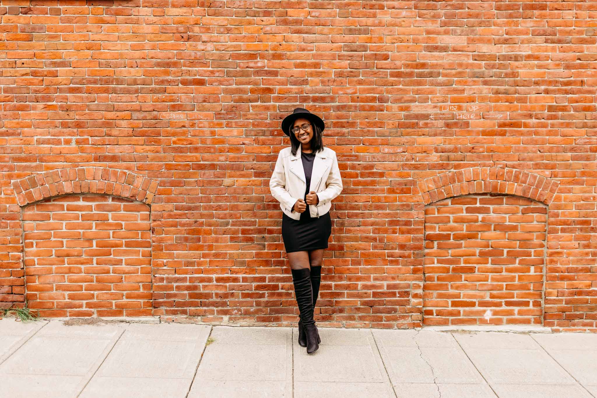 Fall senior portrait of a young black woman wearing boots and a fedora leaning against a brick wall in a city in Massachusetts