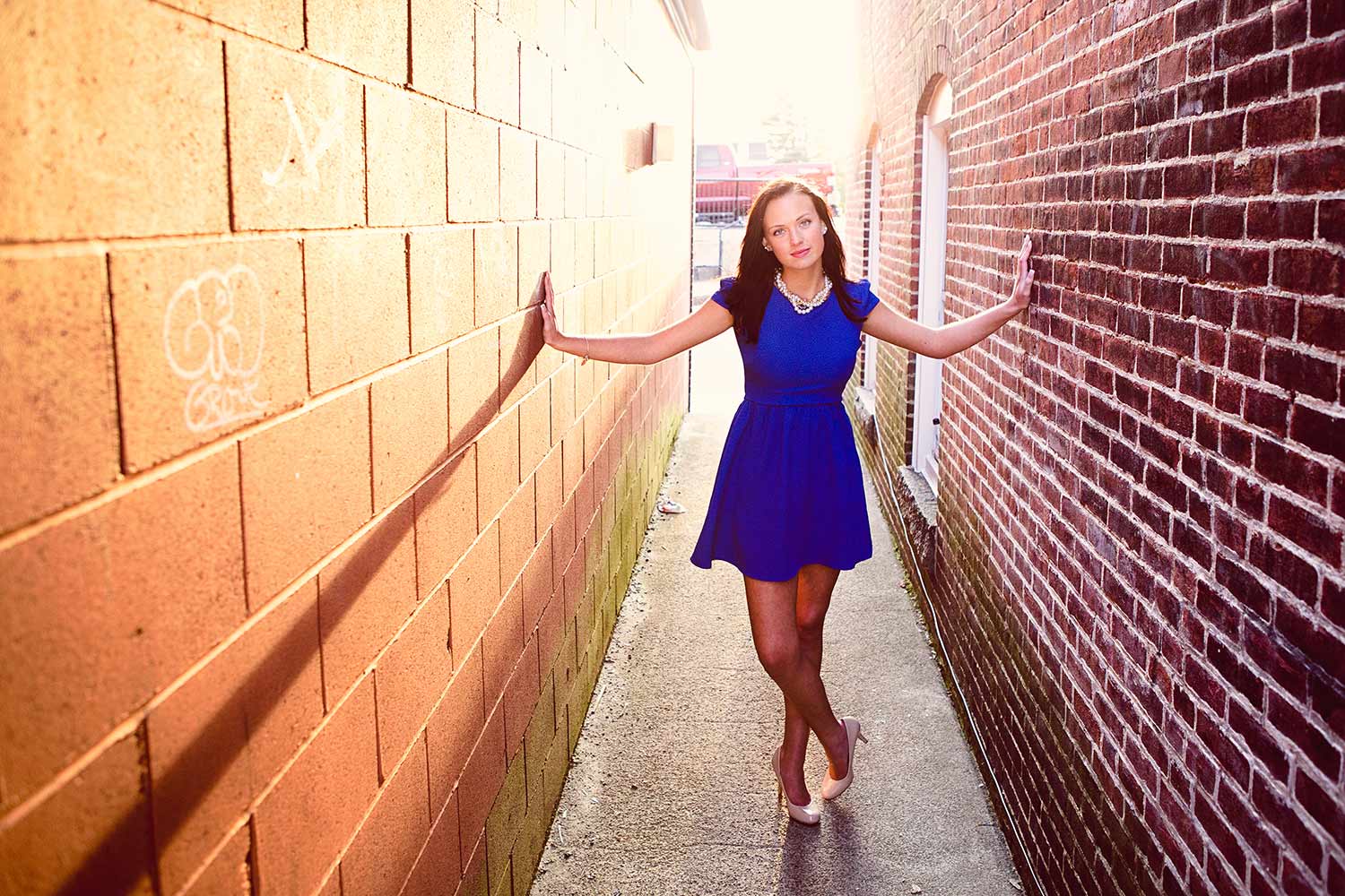 Summer Senior portrait of a young woman in a blue dress with pearls and heels standing in an alleyway in Massachusetts at sunset