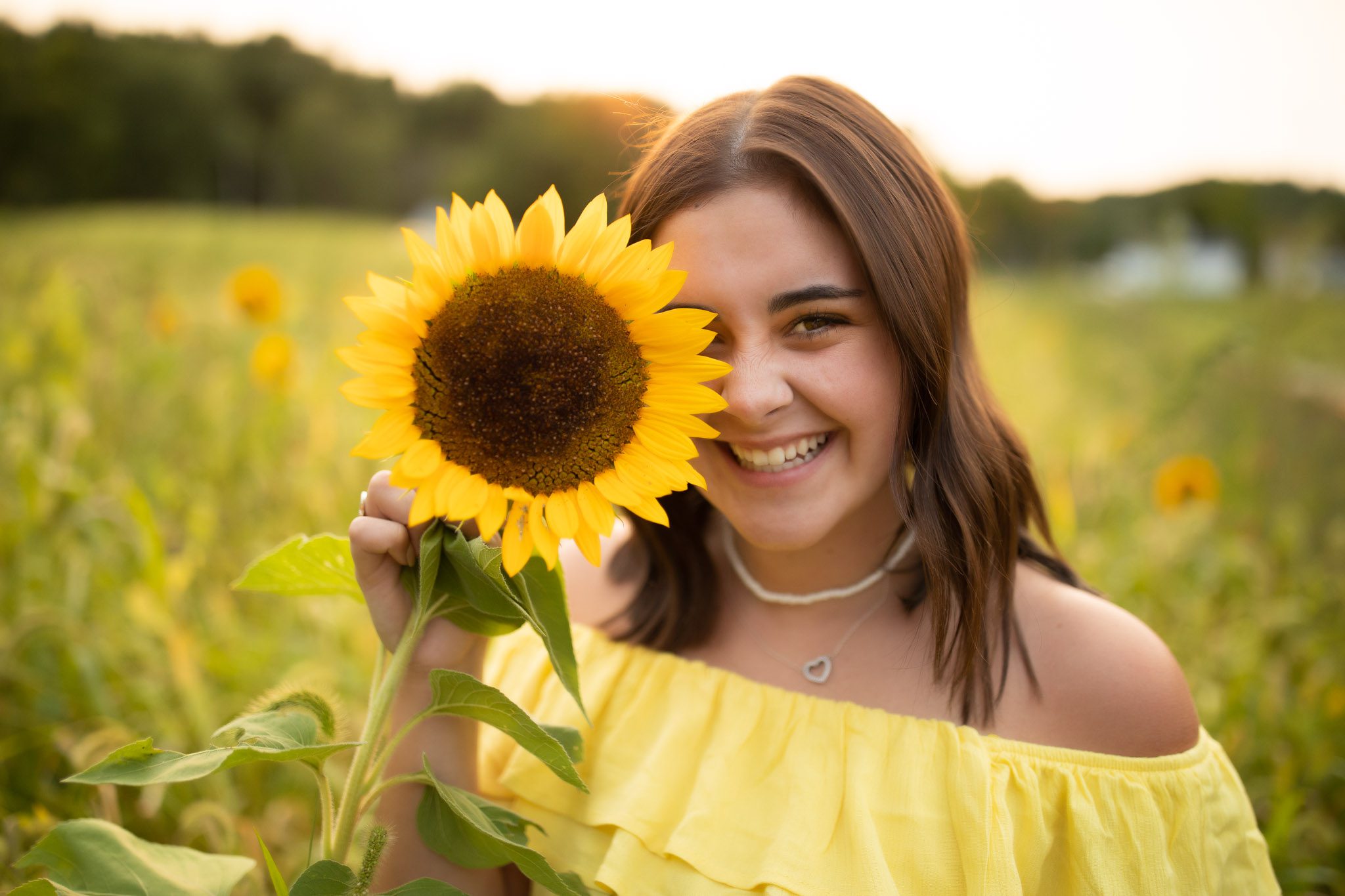 high school summer senior portrait of a young woman in a yellow dress standing in a field of sunflowers holding a sunflower up to her face in Massachusetts