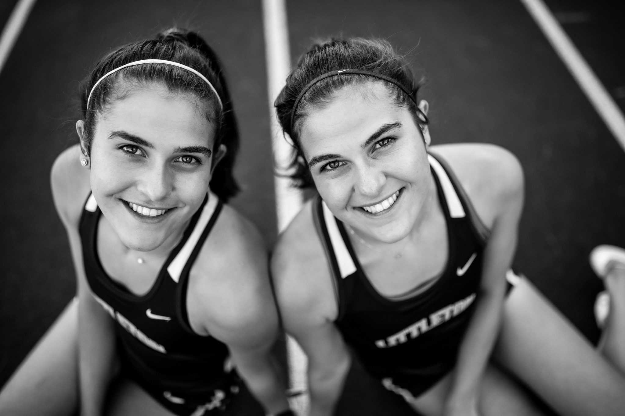 Black and white High school senior portrait of twin girls in racing uniforms sitting on a running track in Littleton, Massachusetts