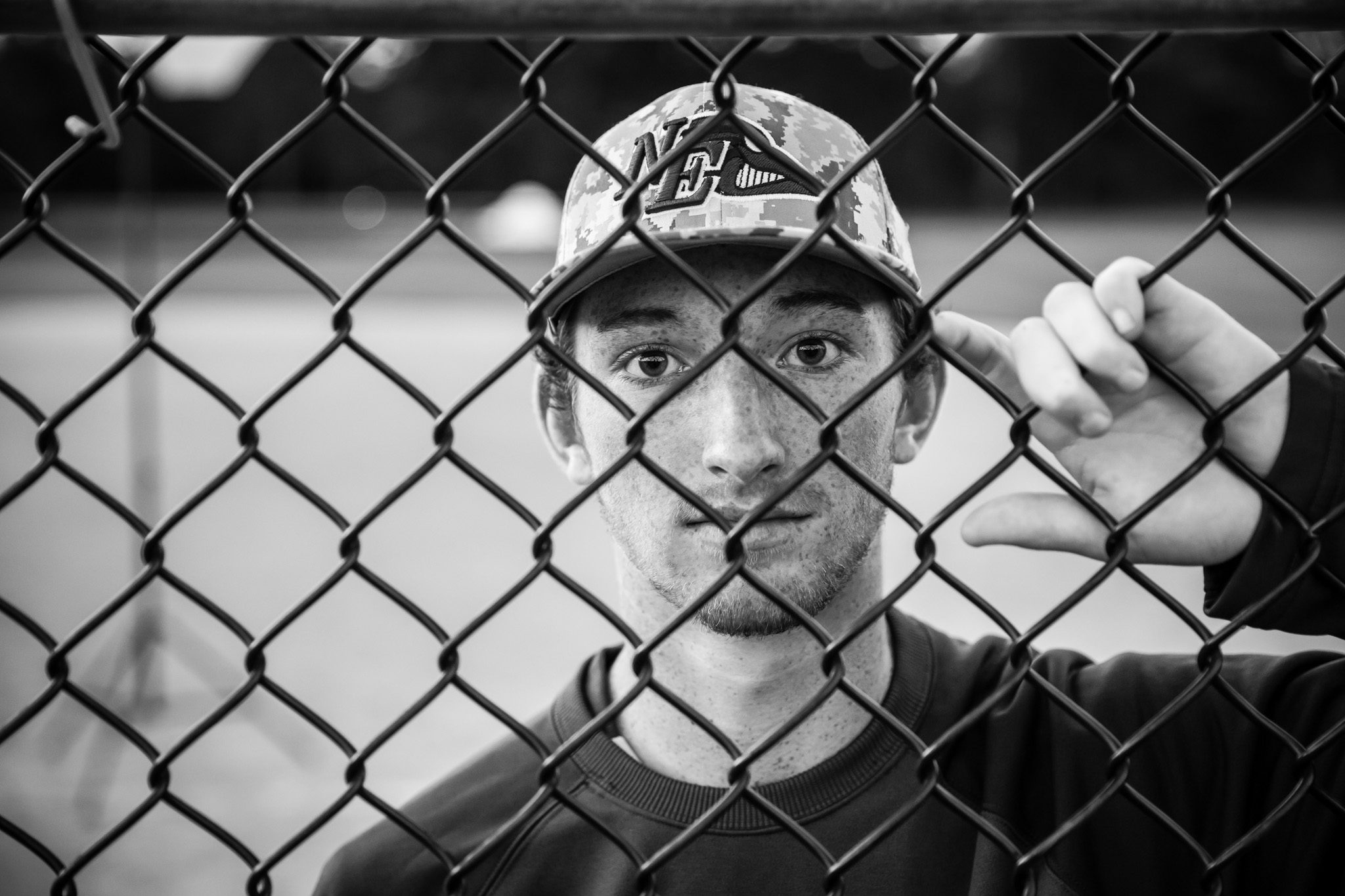 Black and white senior portrait of a boy with freckles in a baseball cap looking through a fence in Massachusetts
