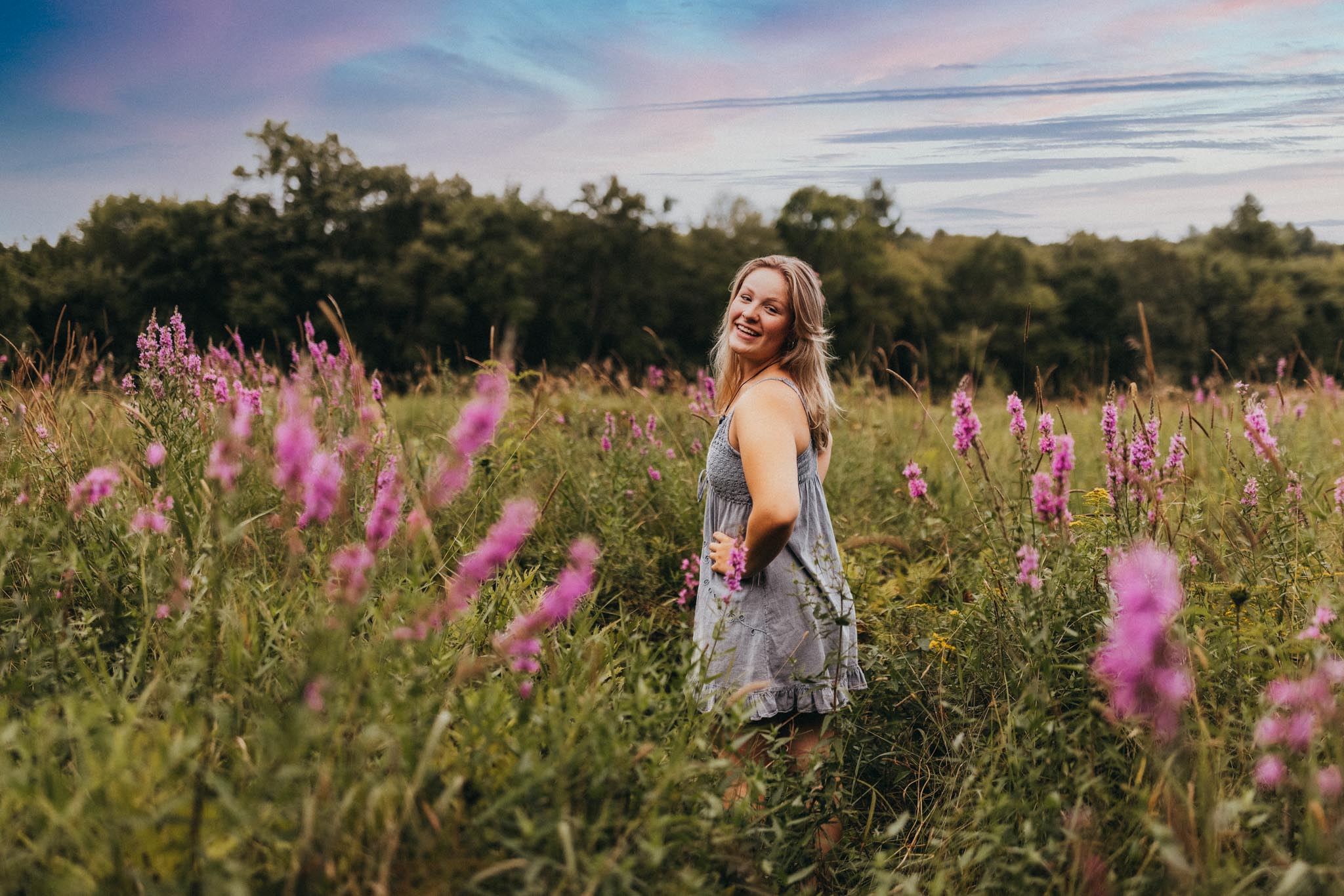 Summer senior portrait of a young woman wearing a sundress standing in a field of purple flowers at sunset in Massachuetts