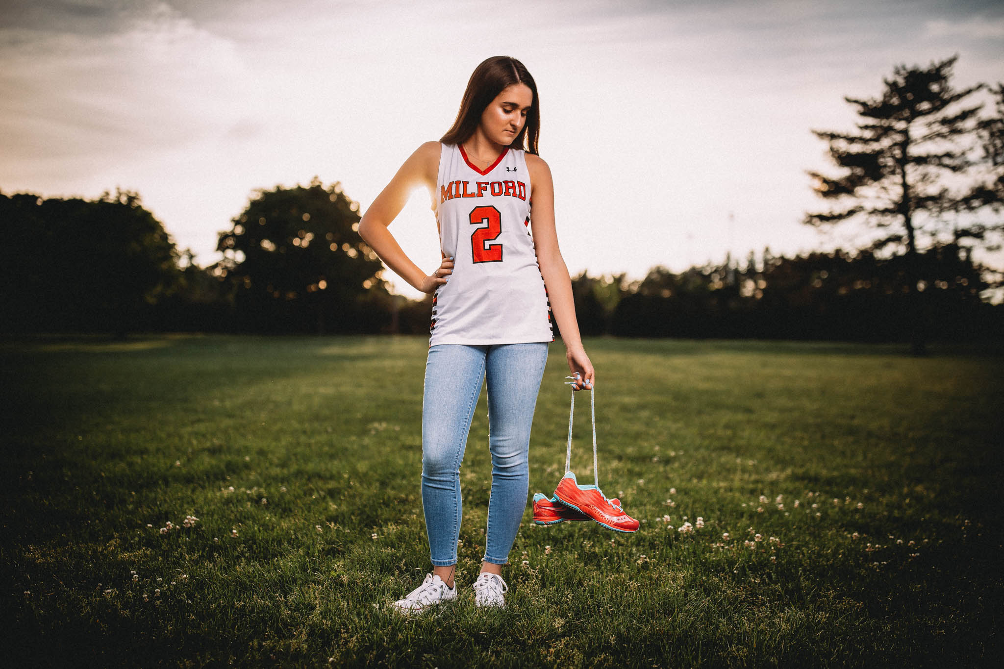 High school senior portrait of a young woman wearing a Milford jersey and holding running shoes standing in a field in Massachusetts at sunset